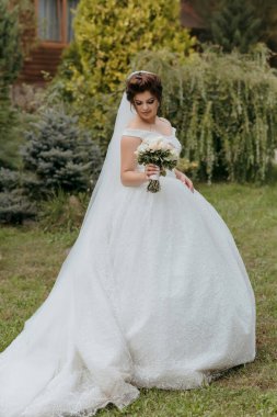 Beyaz gelinlikli bir kadın elinde bir buket çiçekle bir tarlada yürüyor. Sahne sakin ve huzurlu, kadın kameraya bakıyor.