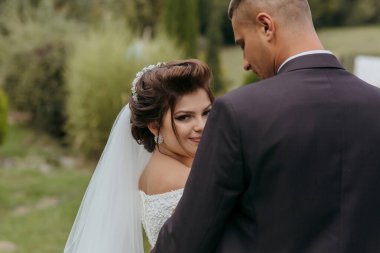 Gelin ve damat birlikte bir tarlada duruyorlar, gelin beyaz bir elbise giyiyor ve damat takım elbise giyiyor. Gelin gülümsüyor ve kameraya bakıyor, damat ise ona bakıyor.