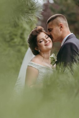 Bir gelin ve damat ormanda öpüşüyor. Gelin beyaz bir peçe takıyor ve damat da takım elbise giyiyor.