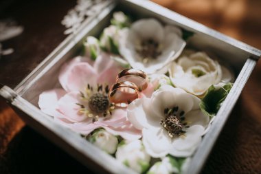 Üzerinde iki altın alyans olan beyaz ve pembe bir çiçek aranjmanı. Yüzükler çiçeklerin üzerine yerleştiriliyor, romantik ve zarif bir atmosfer yaratıyor.