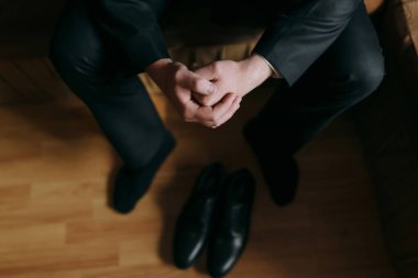 Bir adam ellerini kucağına sıkıştırmış bir kanepede oturuyor. Siyah ayakkabı ve siyah pantolon giyiyor.