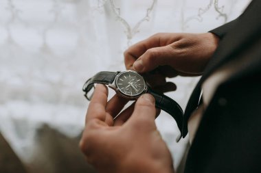 Bir adam elinde bir saat tutuyor. Saat siyah ve gümüş bir şeridi var. Adam takım elbise ve kravat takıyor.