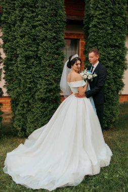 Gelin ve damat çalıların önünde duruyorlar, gelin beyaz bir elbise giyiyor ve damat takım elbise giyiyor.