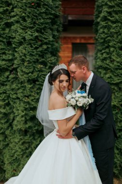 Bir gelin ve damat yeşil bir çalılığın önünde sarılıyorlar. Damat takım elbise giyerken gelin beyaz bir elbise ve peçe takıyor. Bir buket çiçek ve çiçek tutuyorlar.