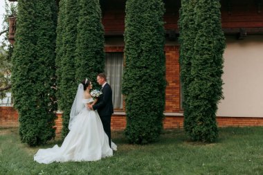 Gelin ve damat bir sıra ağacın önünde durur, gelin beyaz bir elbise giyer ve damat takım elbise giyer.