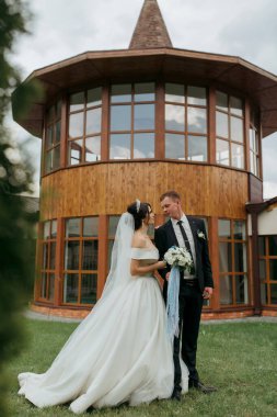 Bir gelin ve damat büyük bir binanın önünde kameraya gülümsüyor. Gelin beyaz bir elbise ve duvak giyerken damat takım elbise ve kravat takıyor. Çift mutlu görünüyor.