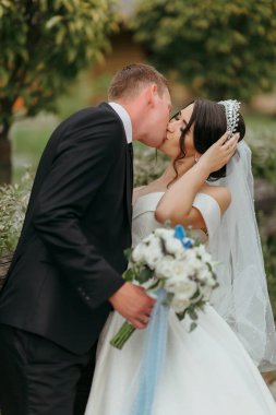 Bir buket çiçeğin önünde bir gelin ve damat öpüşüyor. Gelin beyaz bir elbise ve mavi bir duvak giyiyor. Damat takım elbise ve kravat takıyor. Sahne bir düğün fotoğrafı.