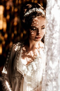 Beyaz elbiseli bir kadın pencerenin önünde duruyor, sırtı kameraya dönük. Elbise beyaz ve dantelli. Kadın taç takıyor.