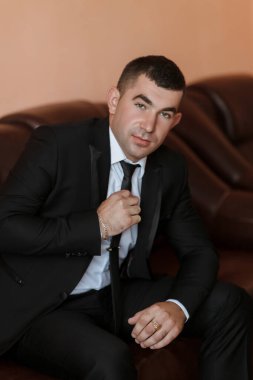 Takım elbiseli ve kravatlı bir adam kanepede oturuyor. Kravatını düzeltiyor ve profesyonel bir ortamda.