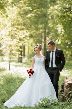 Gelin ve damat ormanda, gelinin elinde bir buket kırmızı gülle dikiliyorlar. Sahne romantik ve huzurlu, çünkü çiftler doğada birlikte vakit geçirmeyi seviyorlar.