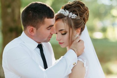 Bir gelin ve damat parkta birbirini kucaklıyor. Gelin beyaz bir elbise ve taç giyiyor.