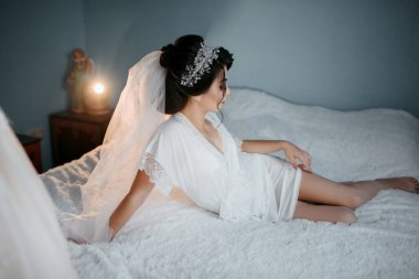 Beyaz elbiseli bir kadın yatakta yatıyor. Duvak ve taç takıyor.