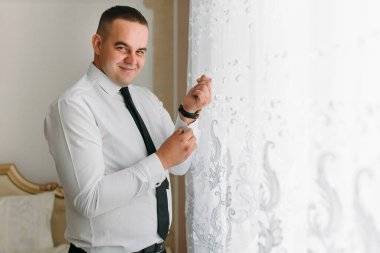 Beyaz gömlekli ve siyah kravatlı bir adam pencerenin önünde giyiniyor. Gülümsüyor ve keyfi yerinde.