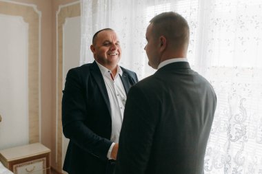 İki adam pencerenin önünde duruyor, biri takım elbise giyiyor ve diğeri de kravat takıyor. Gülümsüyorlar ve el sıkışıyorlar.