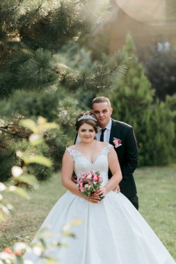 Bir gelin ve damat tarlada fotoğraf çektirmek için poz veriyorlar. Gelin beyaz bir elbise giyiyor ve damat takım elbise giyerken elinde bir buket tutuyor. Sahne huzurlu ve romantik.