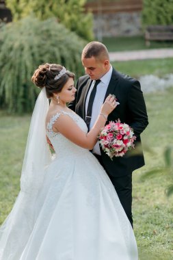 Gelin ve damat çimenli bir tarlada, gelin beyaz bir elbise ve damat siyah bir takım elbise giyiyor. Gelin bir buket çiçek tutuyor ve damat onun eline dokunuyor.