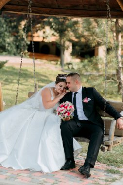 Gelin ve damat salıncakta oturuyor, gelin de elinde buketle. Sahne romantik ve mutlu.