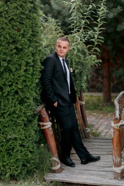 Takım elbiseli bir adam çalılığın önünde duruyor. O bir kravat ve siyah bir ceket giyiyor.