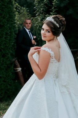 Bir gelin ve damat bir çalılığın önünde fotoğraf için poz veriyorlar. Damat takım elbise giyerken gelin beyaz bir elbise ve taç giyiyor. Sahne mutlu ve kutlama dolu.