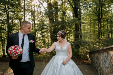 Bir gelin ve damat ormanda bir patikada yürüyorlar. Gelin beyaz bir elbise giyiyor ve damat bir takım elbise giyiyor. Gelin bir buket tutuyor ve damat elini tutuyor.