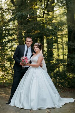 Bir gelin ve damat ormanda fotoğraf çektirmek için poz veriyorlar. Gelin beyaz bir elbise giyiyor ve damat bir takım elbise giyiyor. Ellerinde buket ve buket tutuyorlar.