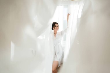 Beyaz elbiseli bir kadın perdenin önünde duruyor. O gülümsüyor ve pencereden dışarı bakıyor. Sıcaklık ve mutluluk kavramı, kadın dışarıdaki manzaranın tadını çıkarırken