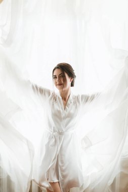 Beyaz cüppeli bir kadın pencerenin önünde, perdeleri açık duruyor. Gülümsüyor ve gün ışığının tadını çıkarıyor. Sıcaklık ve mutluluk kavramı