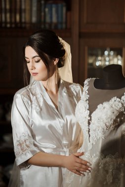 Bir gelin gelinliğin önünde duruyor, göğsüne dayıyor. Elbise beyaz ve dantelli. Gelin beyaz bir cübbe ve duvak giyiyor.