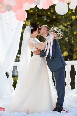 Gelin ve damat pembe kemerin önünde öpüşürler. Gelin beyaz bir elbise giyiyor ve damat mavi bir takım giyiyor.