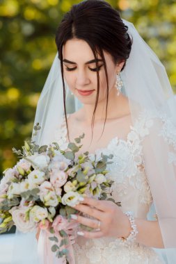 Beyaz gelinlik giyen bir kadın elinde bir buket çiçek tutuyor. Saçları topuz gibi ve peçe takıyor. Buket pembe çiçeklerle dolu, sahneyi romantikleştiriyor.