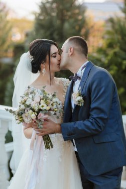 Beyaz bir kemerin önünde bir gelin ve damat öpüşüyor. Gelin bir buket çiçek tutuyor ve damat mavi bir takım elbise giyiyor.