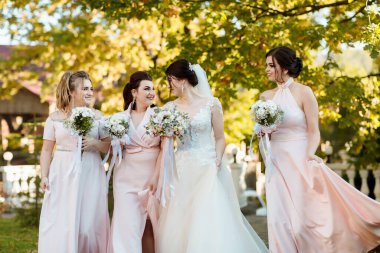 Bir grup kadın birlikte yürüyor, içlerinden biri gelinlik giyiyor. Gelin gülümsüyor ve diğer kadınlar da gülümsüyor. Mutlu ve neşeli bir ortam yaratıyorlar.