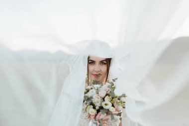 Beyaz bir peçenin önünde elinde bir buket çiçekle bir gelin duruyor. Sahne açık mavi gökyüzü olan bir tarlada geçiyor.