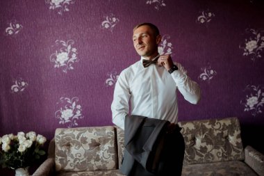 Takım elbiseli ve papyonlu bir adam kanepenin önünde duruyor. Kravatını düzeltiyor ve kameraya bakıyor. Adam takım elbise ve kravat giydiği için resmiyet ve zarafet kavramı.