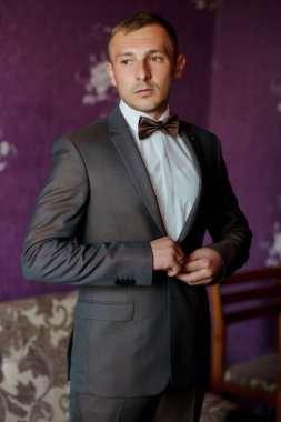 Takım elbiseli ve kravatlı bir adam mor bir duvarın önünde duruyor. Kravatını düzeltiyor ve kameraya bakıyor.