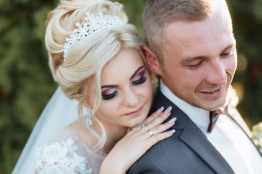 Bir gelin ve damat düğün fotoğrafında birbirlerine sarılıyorlar. Gelin taç takıyor ve damat da takım elbise giyiyor. Sahne mutlu ve romantik.