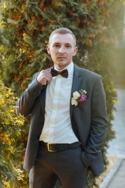 Takım elbiseli ve kravatlı bir adam resim için poz veriyor. Papyon ve yaka çiçeği takıyor. Resmin resmi ve zarif bir havası var.