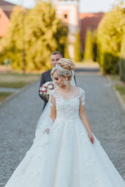 Gelin ve damat kaldırım taşı bir caddede duruyorlar, gelin beyaz bir elbise giyiyor ve damat takım elbise giyiyor. Gelin elinde bir buket çiçek tutuyor.