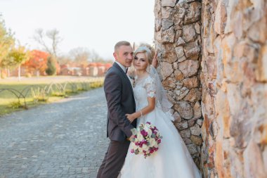 Bir gelin ve damat taş bir duvarın önünde poz veriyorlar. Gelin beyaz bir elbise giyiyor ve damat bir takım elbise giyiyor. Sahne mutlu ve romantik.