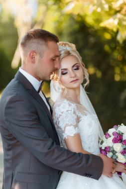 Bir gelin ve damat parkta birbirini kucaklıyor. Gelin beyaz bir elbise giyiyor ve damat bir takım elbise giyiyor. Bir buket çiçek ve bir vazo tutuyorlar. Sahne romantik ve mutlu.
