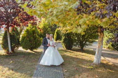 Bir gelin ve damat, yaprakları renk değiştiren bir ağacın önünde duruyorlar. El ele tutuşup gülümsüyorlar.