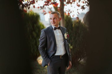 Takım elbiseli ve papyonlu bir adam bir ağacın önünde duruyor. O, beyaz bir gömlek ve yakasında pembe bir çiçek giyiyor.