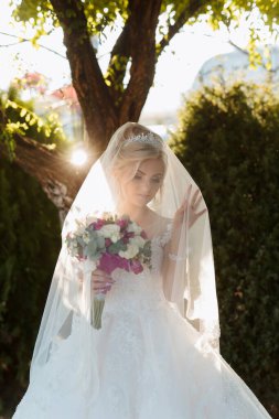 Bir gelin peçe takıyor ve elinde bir buket çiçek tutuyor. Bir ağacın önünde duruyor.