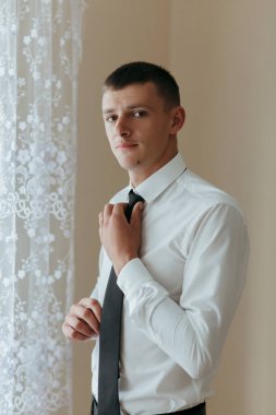 Beyaz gömlekli ve siyah kravatlı bir adam pencerenin önünde duruyor. Kravatını düzeltiyor ve kameraya bakıyor. Adam takım elbise giydiği için resmiyet ve profesyonellik kavramı.