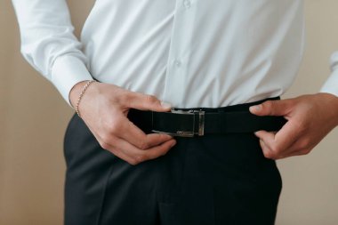 Bir adam beyaz gömlek ve siyah pantolon giyiyor. O, kemerini ayarlıyor. Formalite kavramı ve detaylara dikkat