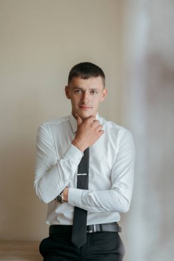 Beyaz gömlekli ve siyah kravatlı bir adam bir duvarın önünde duruyor. Kameraya bakıyor ve derin düşüncelere dalmış.