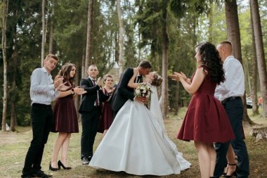 Bir gelin ve damat bir grup insanın önünde öpüşüyor. Gelin beyaz bir elbise giyiyor ve damat bir takım elbise giyiyor. Çevrelerindeki insanlar alkış ve tezahürat yapıyor.