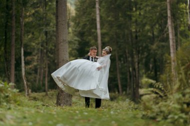 Gelin ve damat ormanda duruyorlar, gelin elinde bir buket çiçekle. Sahne romantik ve huzurlu, çiftin doğa ile çevrili özel günlerinin tadını çıkardığı gibi.