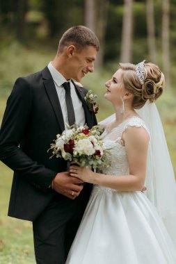 Gelin ve damat tarlada duruyorlar, gelin beyaz bir elbise giyiyor ve damat siyah bir takım elbise giyiyor.