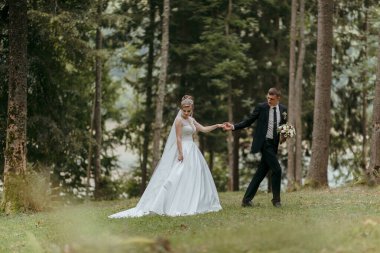Bir gelin ve damat el ele bir ormanda yürüyorlar. Gelin beyaz bir elbise giyiyor ve damat bir takım elbise giyiyor. Sahne romantik ve huzurlu.